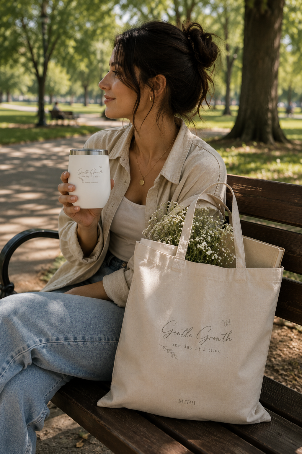 Woman relaxing on a park bench holding the Gentle Growth insulated tumbler with a matching tote bag, enjoying a peaceful outdoor moment.